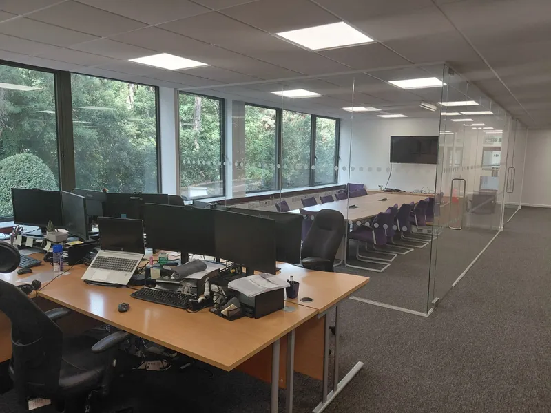 Open-plan office desks beside a glass-walled meeting room and carpeted walkway